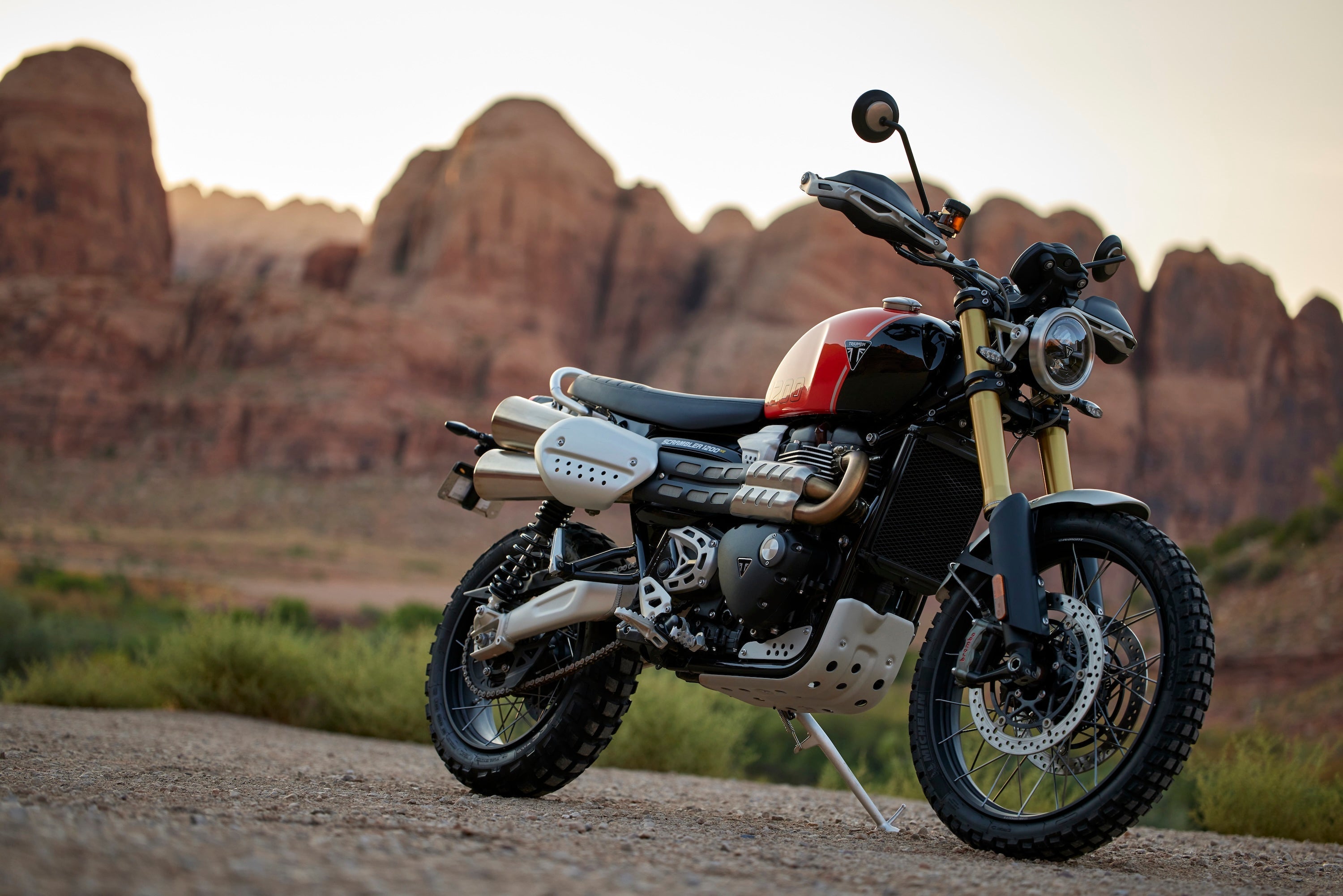 A 2024 Triumph Scrambler 1200 XE in Baja Orange and Phantom Black, parked on a dirt trail with red rock mountains in the background at sunset.