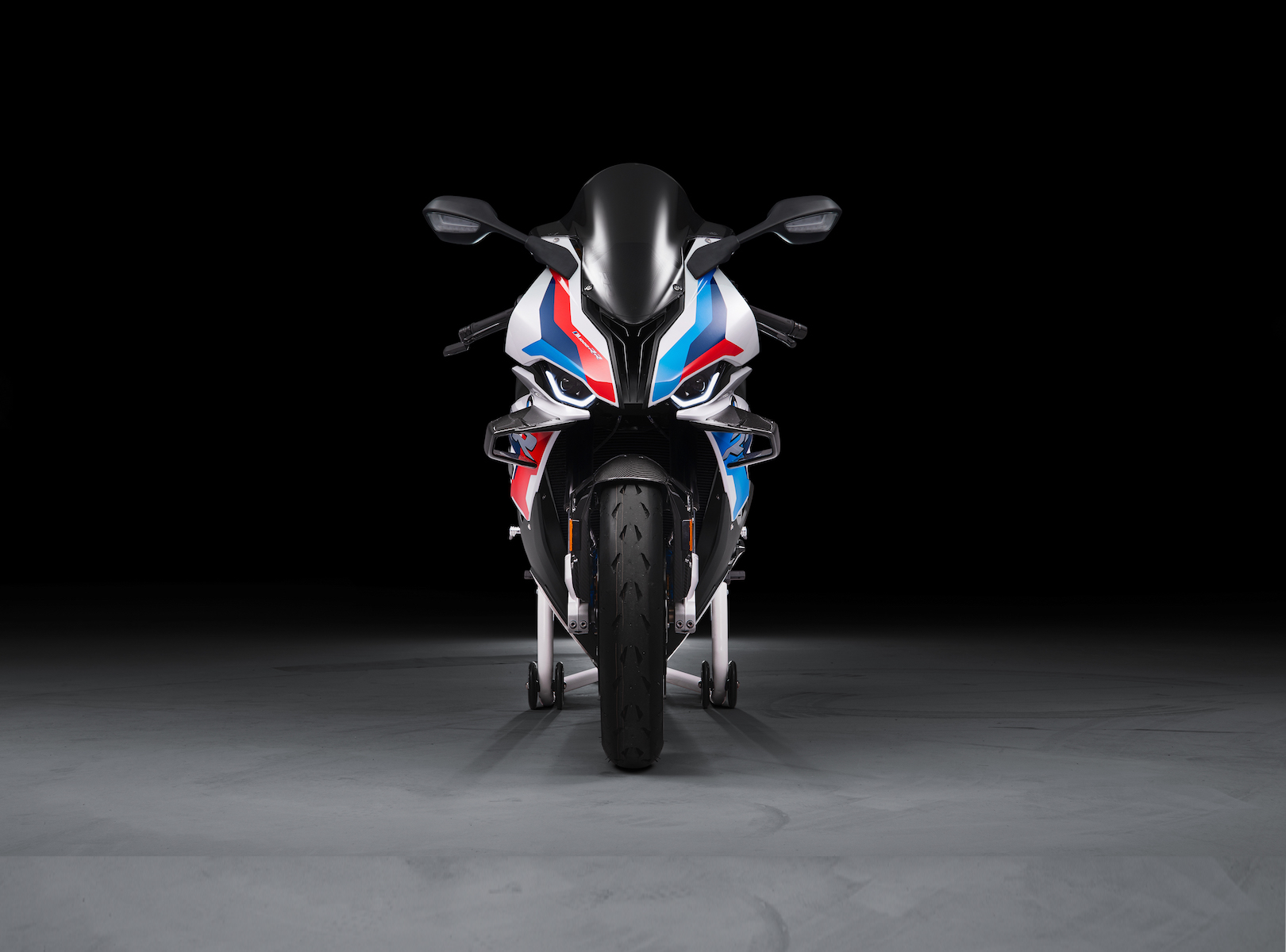 Front view of a BMW M 1000 RR superbike in white, blue, and red M Motorsport livery on a paddock stand in a studio setting.