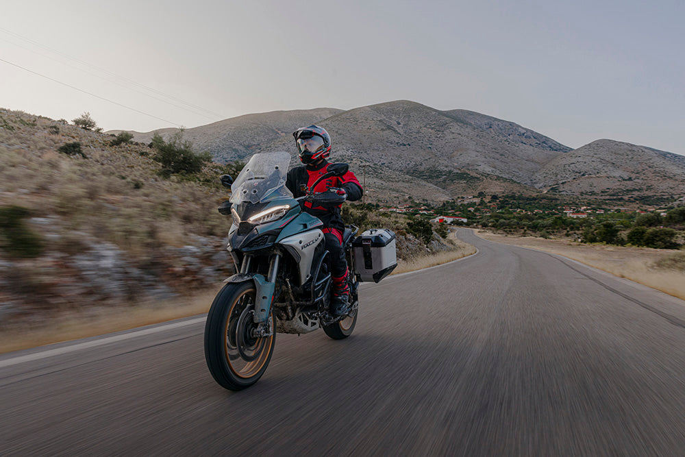 Adventure motorcycle rider touring on a mountain road with side panniers and full riding gear.