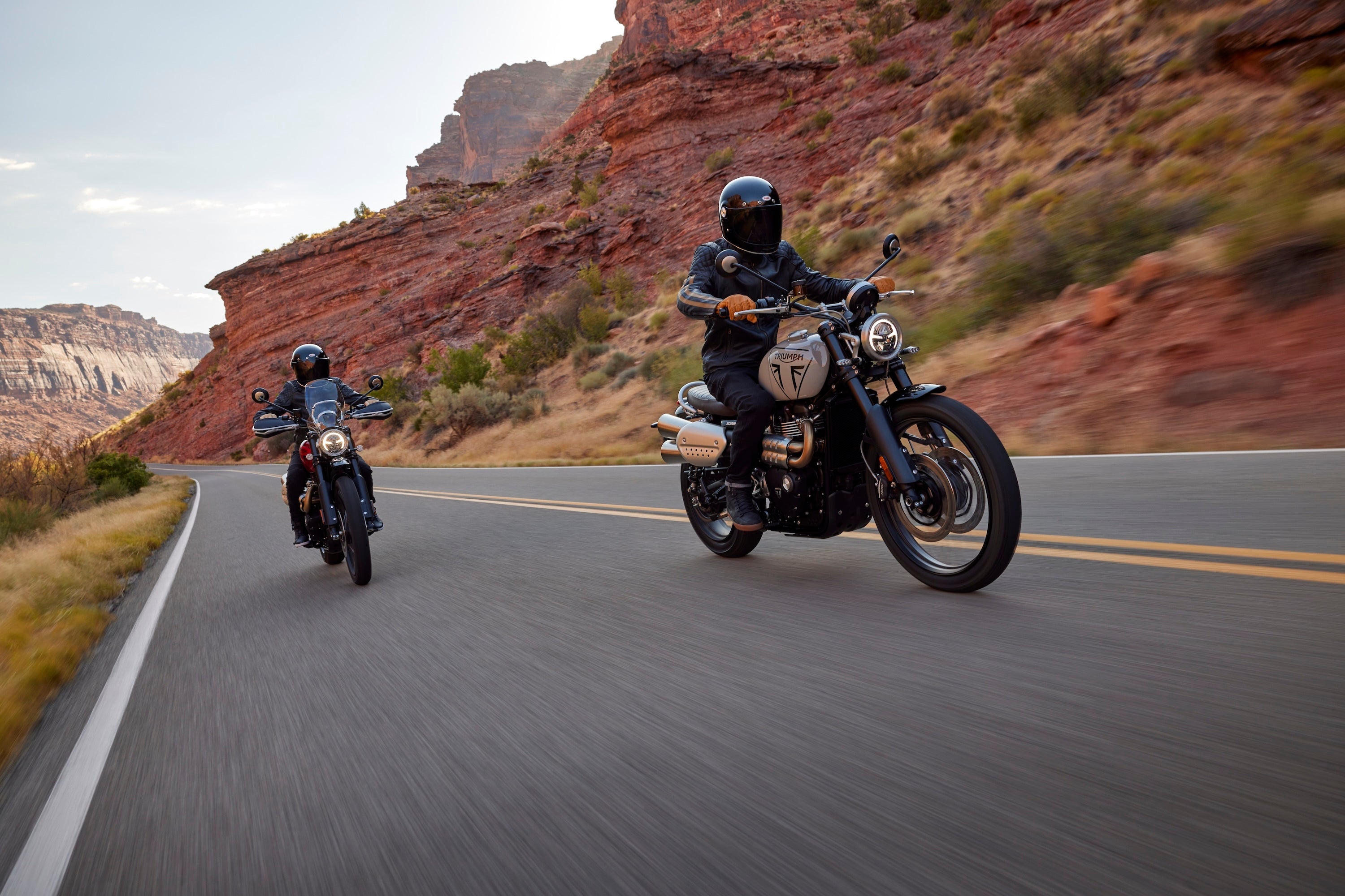 Two motorcyclists riding on a desert road with rocky cliffs in the background