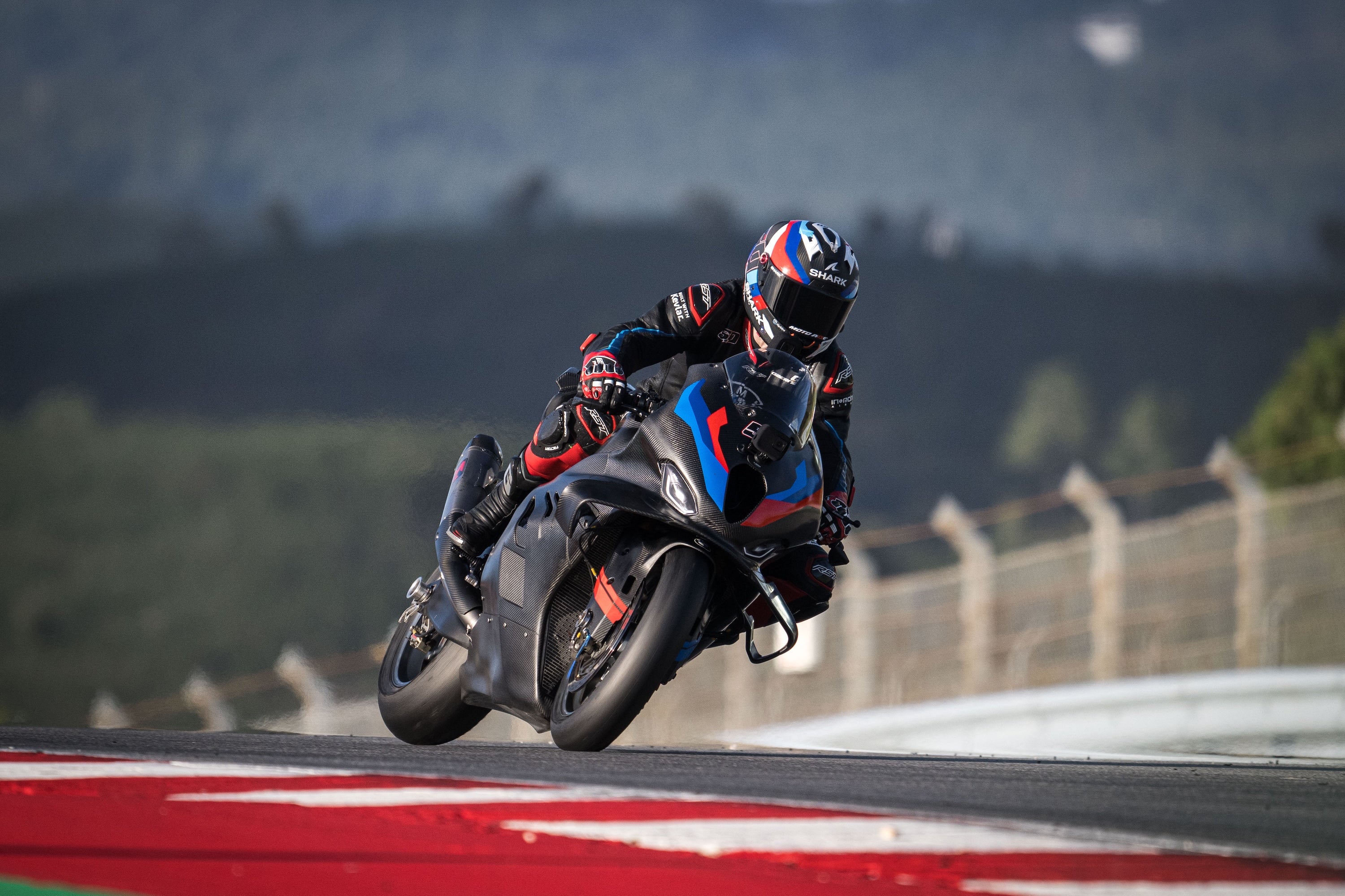 Front-side view of a BMW M Motorsport racing motorcycle cornering on an asphalt track with a blurred green hillside in the background.