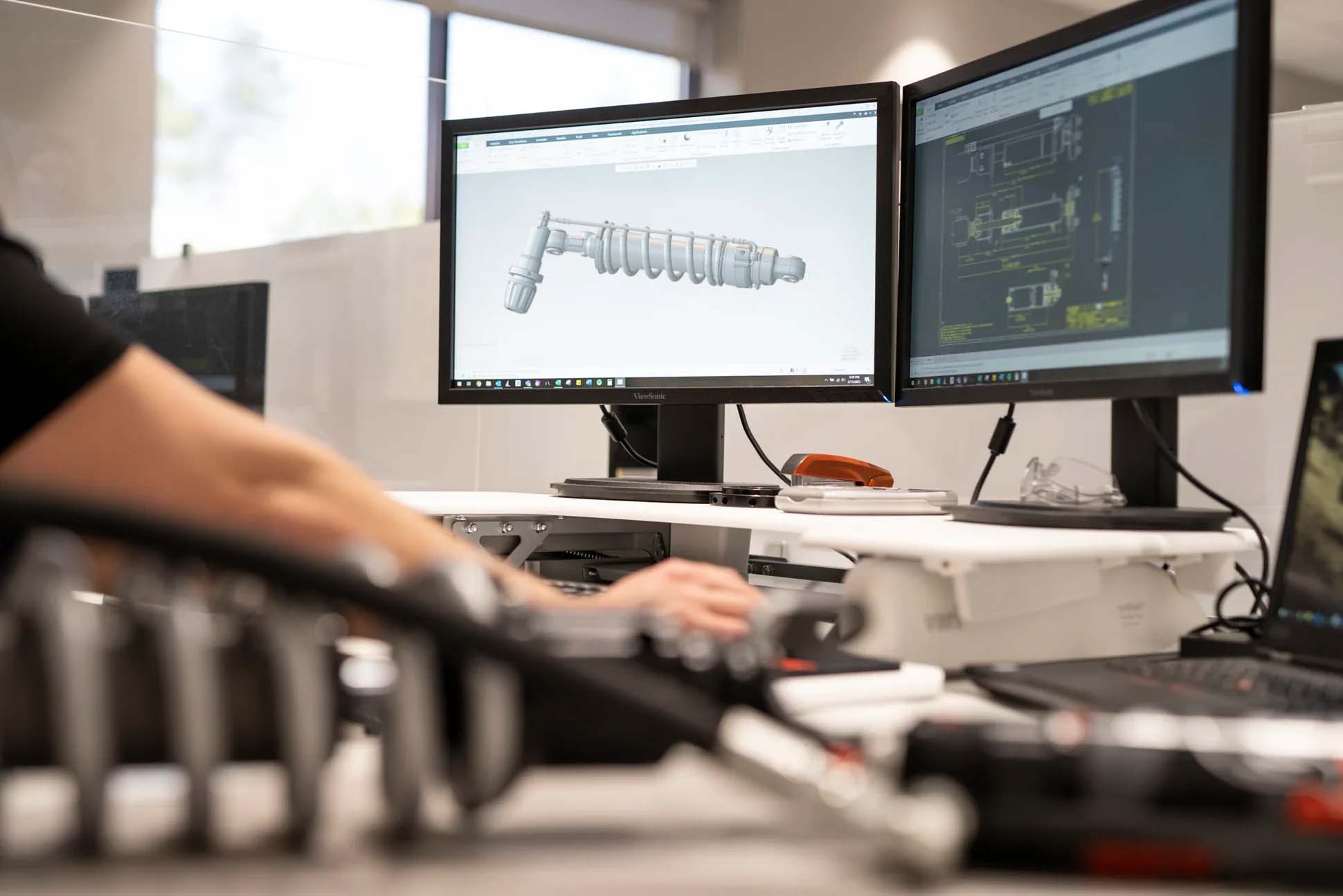 Image DescriptionAn engineer works at a modern dual-monitor workstation in a bright office, focusing on the mechanical design of a motorcycle shock absorber.