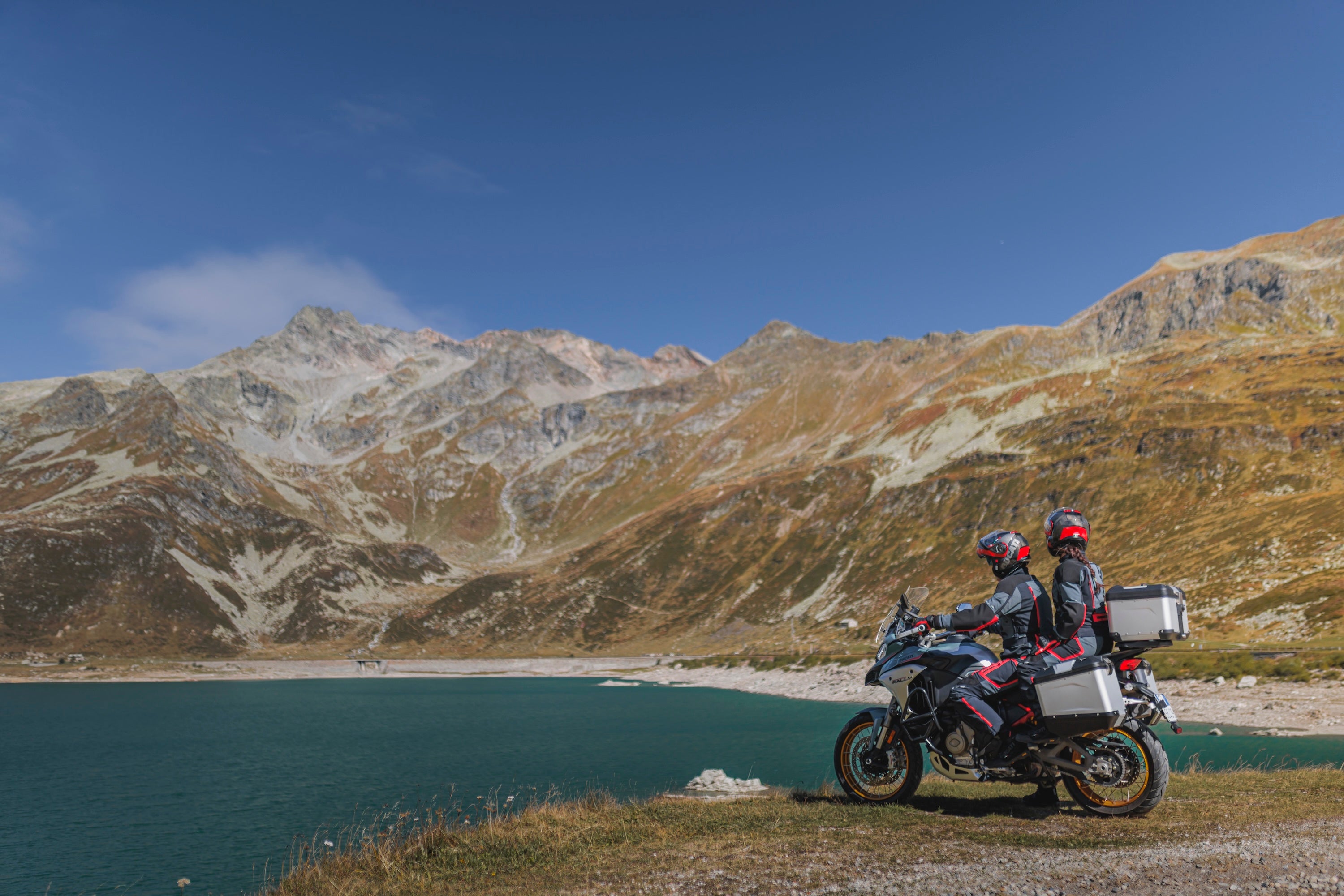 Two people on a Ducati Multistrada adventure motorcycle with silver panniers, parked on a gravel path overlooking a turquoise alpine lake and rugged mountains.