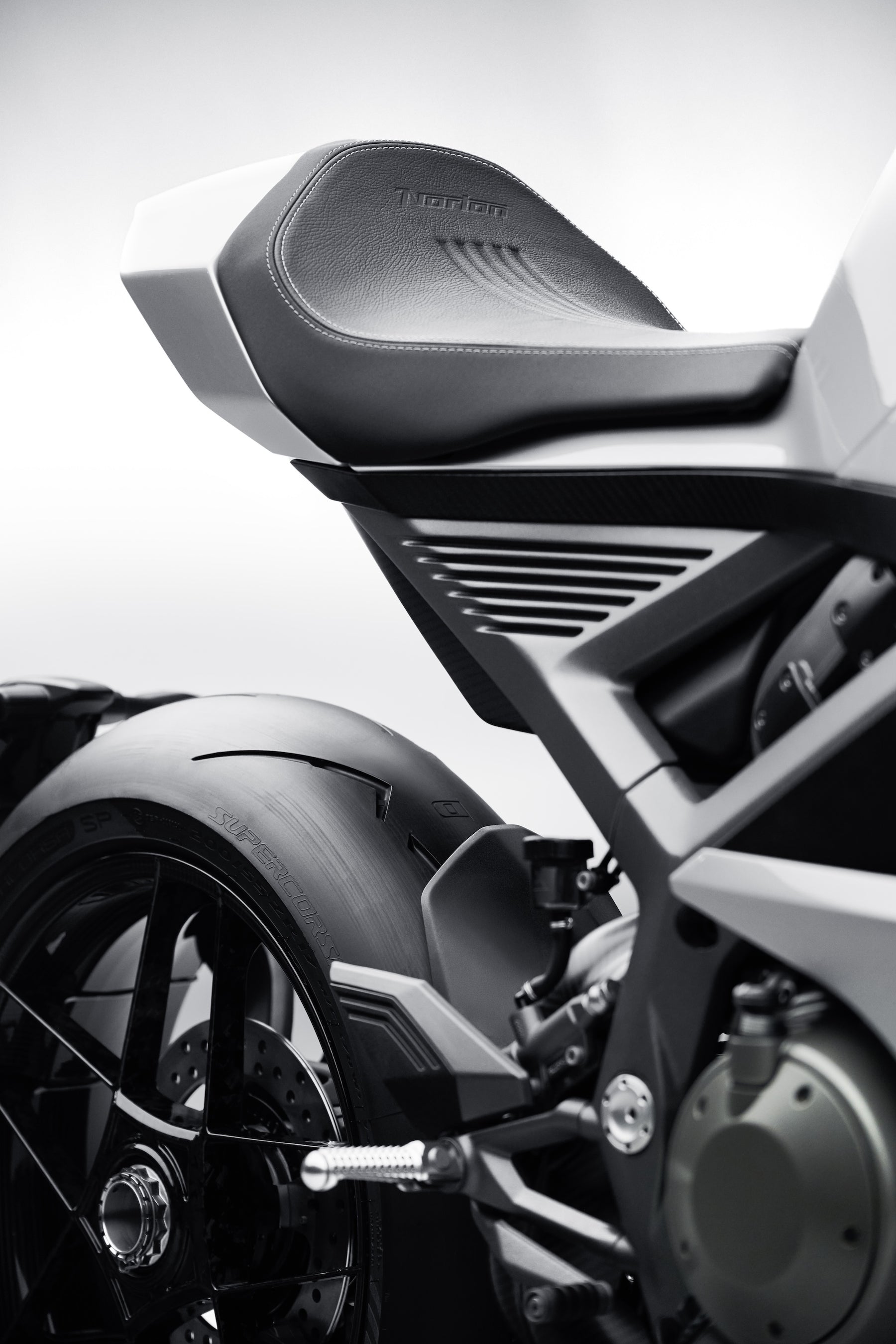 A close-up of a silver motorcycle's rear seat and tire, showing fine stitching on the black leather and the word Norton pressed into the material.