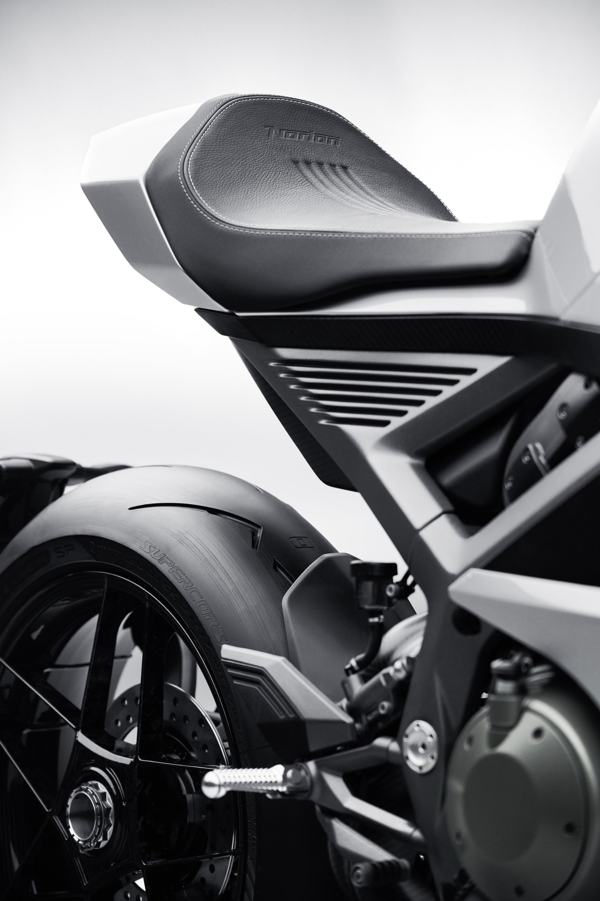 A close-up of a silver motorcycle's rear seat and tire, showing fine stitching on the black leather and the word Norton pressed into the material.
