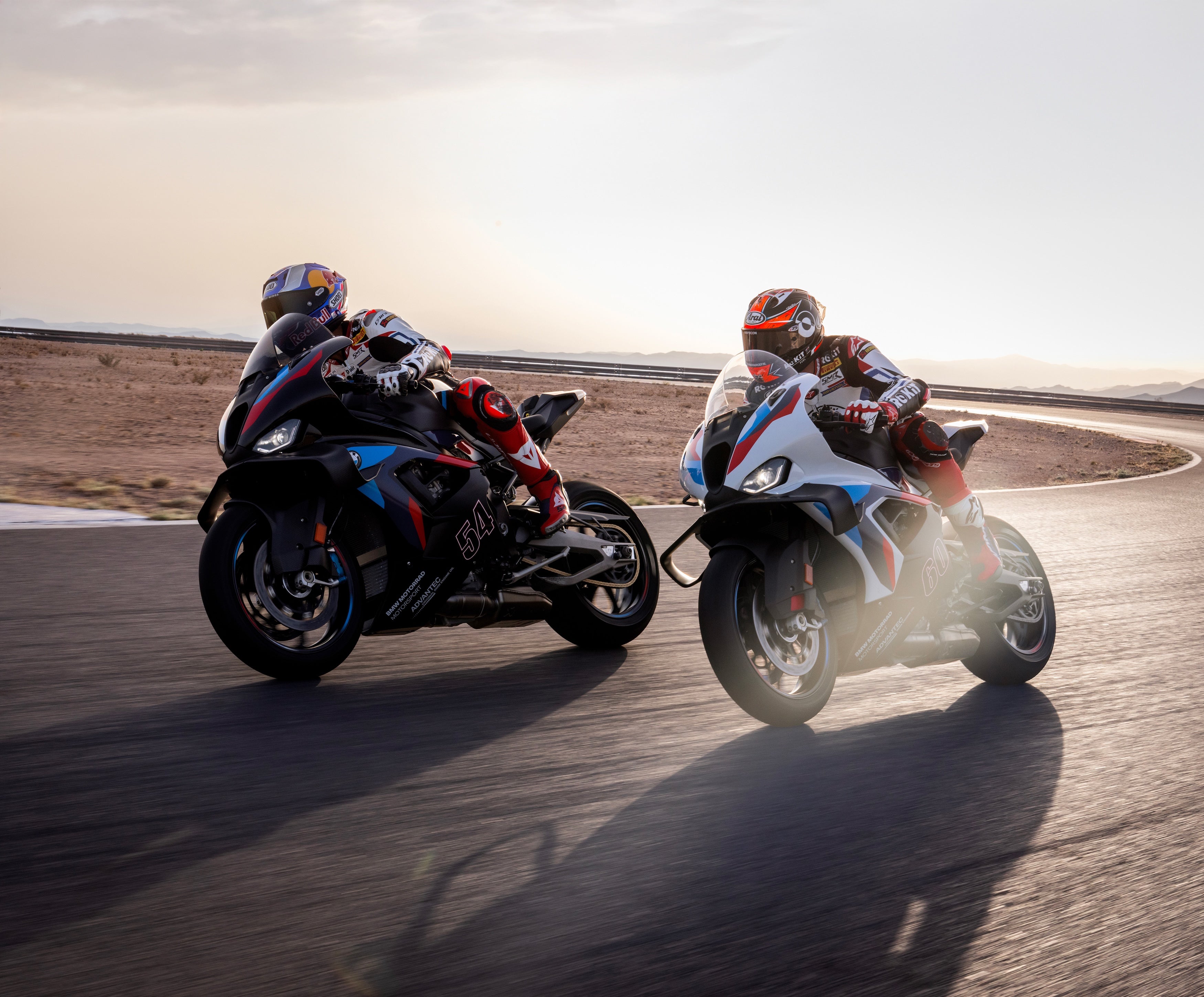 Two BMW M 1000 RR superbikes racing side by side on a desert racetrack at sunset, showcasing high-performance suspension and track-ready handling.