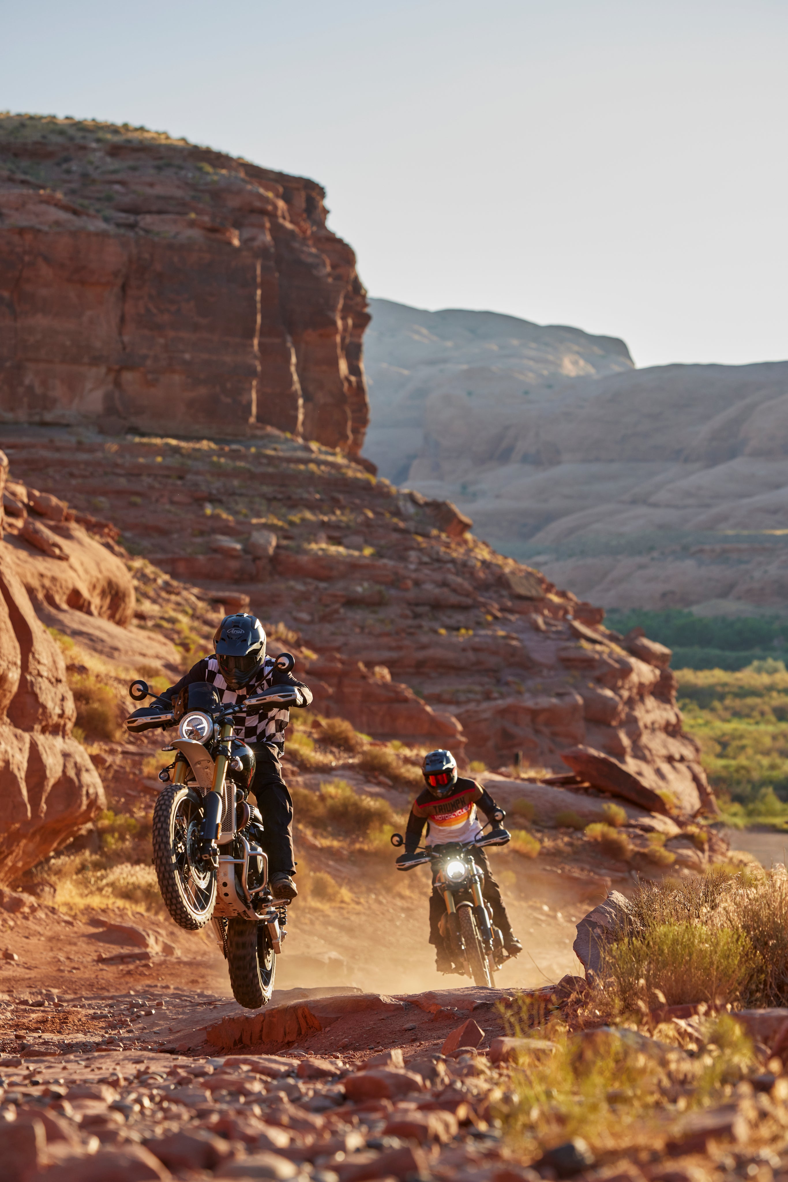 Two adventure motorcyclists riding off-road through a red rock desert trail at sunset.