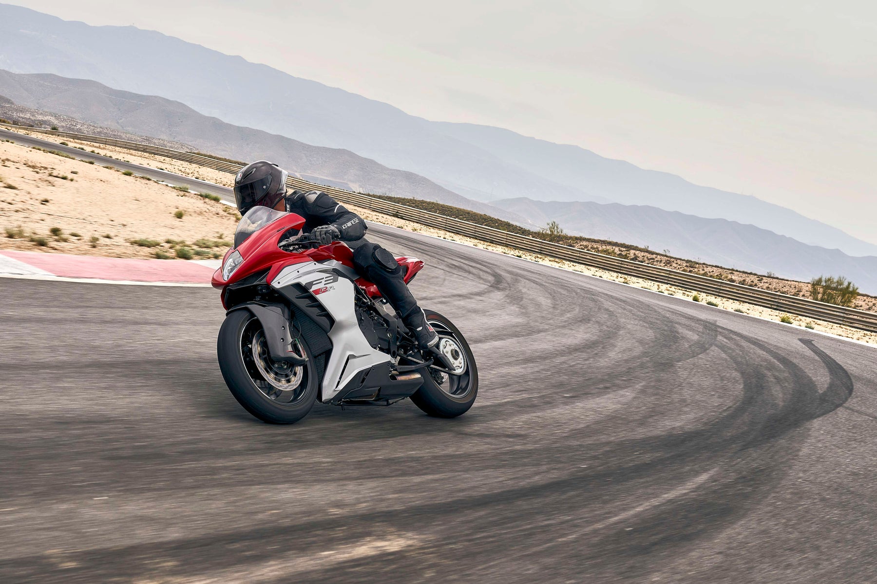 Red sport motorcycle leaning into a high-speed corner on a desert racetrack with mountain backdrop, showcasing performance suspension and precision handling.